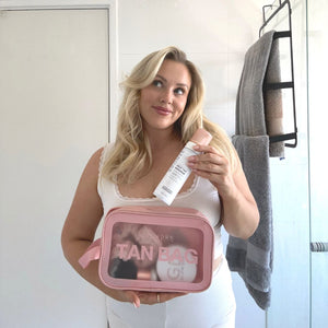 Blonde model wear self tan in white pyjama's looks to the sky with a smile while holding glowdry tan bag cosmetic case make up bag showing clear panel to front and glowdry products inside model is in white bathroom with towels and shower beside her
