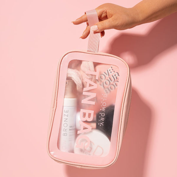 A hand hold Tanbag make up bag by the carry handle pink products can be seen inside as it has clear panel slight shadow to the pin background