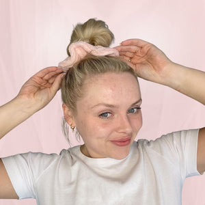 model with blonde hair shows glowdry self tan scrunchie in hair bun hair is tied up and pink background